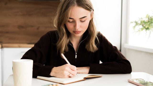 A woman with light brown hair wearing a dark sweater sits at a white table, writing in a notebook with a pen. A white mug, a bowl, and a smartphone are also on the table.