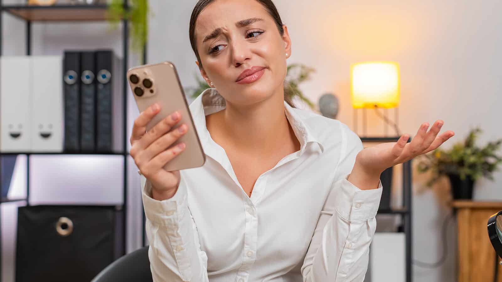 A woman in a white shirt looks at her phone with a confused expression, holding one hand up as if unsure, sitting in an office with shelves and a lamp in the background.