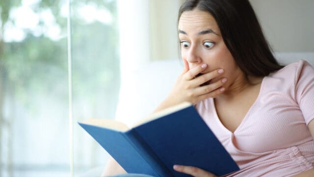 A woman sits indoors, holding a blue book and looking surprised with wide eyes and her hand covering her mouth. Light from a window shines behind her.