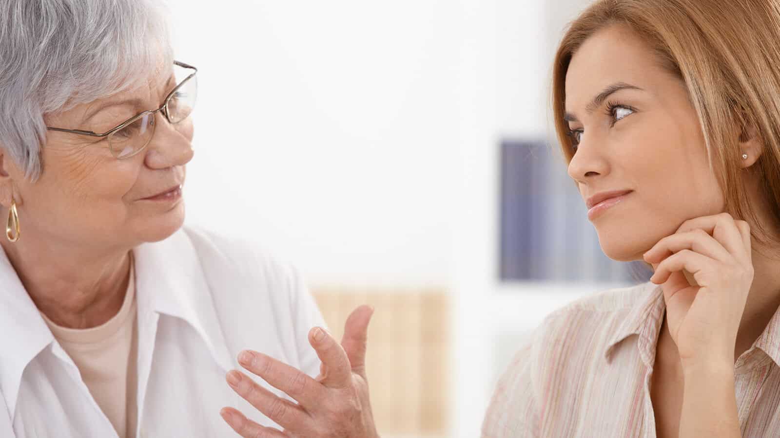 An older woman with gray hair speaks to a younger woman with light brown hair, who listens attentively with her hand on her chin. The two appear to be engaged in a thoughtful conversation indoors, perhaps discussing the psychology of aging and habits for older adults.