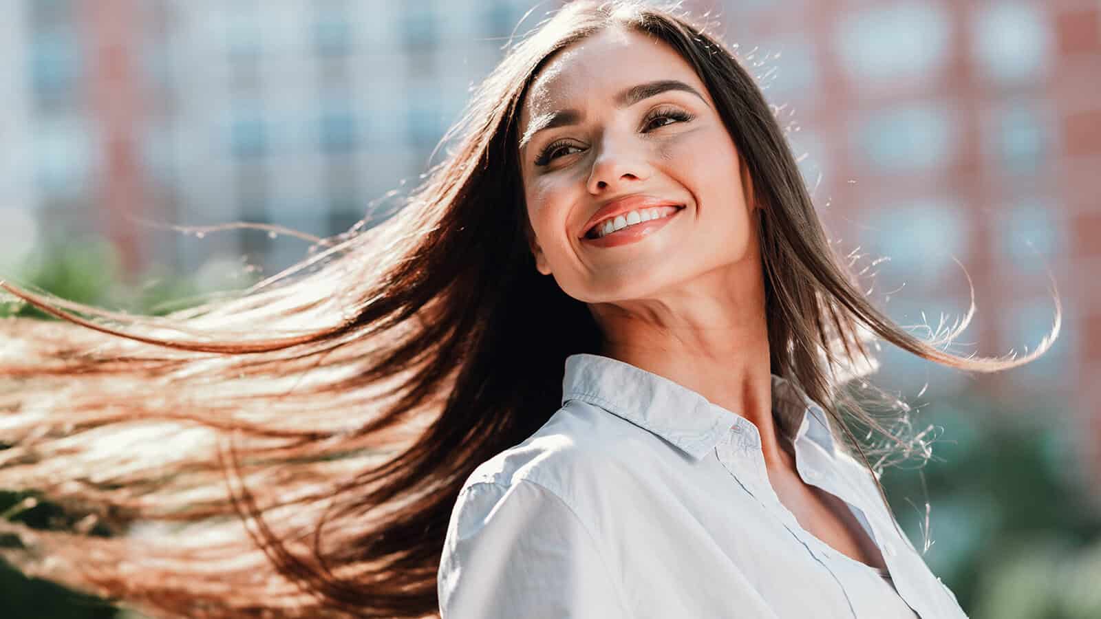 A woman with long brown hair smiles outdoors, wearing a light-colored shirt. The background is blurred with greenery and buildings, suggesting a sunny day.