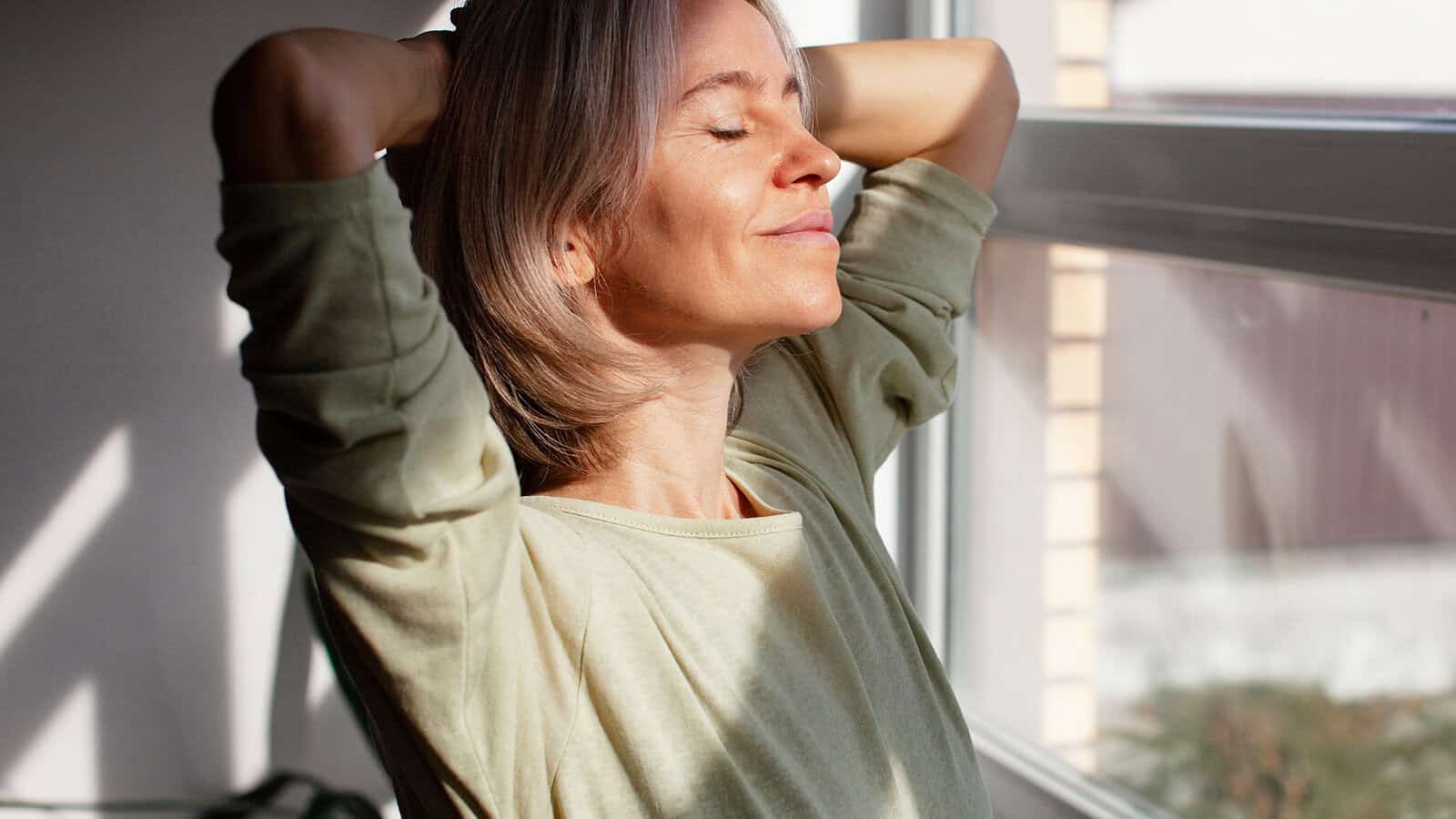A woman with light brown hair relaxes by a window, eyes closed and smiling softly, enjoying the sunlight on her face with her hands resting behind her head.