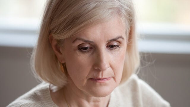 Close-up of a middle-aged woman with light blonde hair and fair skin, looking down with a thoughtful or serious expression, wearing a cream-colored sweater.