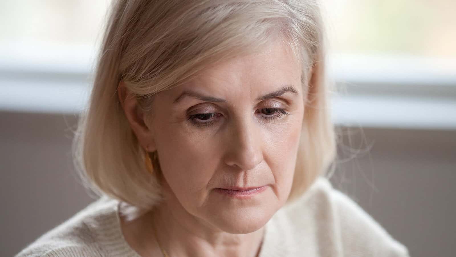 Close-up of a middle-aged woman with light blonde hair and fair skin, looking down with a thoughtful or serious expression, wearing a cream-colored sweater.