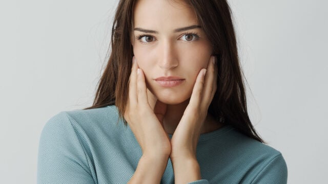A woman with long brown hair and a serious expression gently touches her face with both hands. She is wearing a textured light blue top and is posed against a plain, light background.