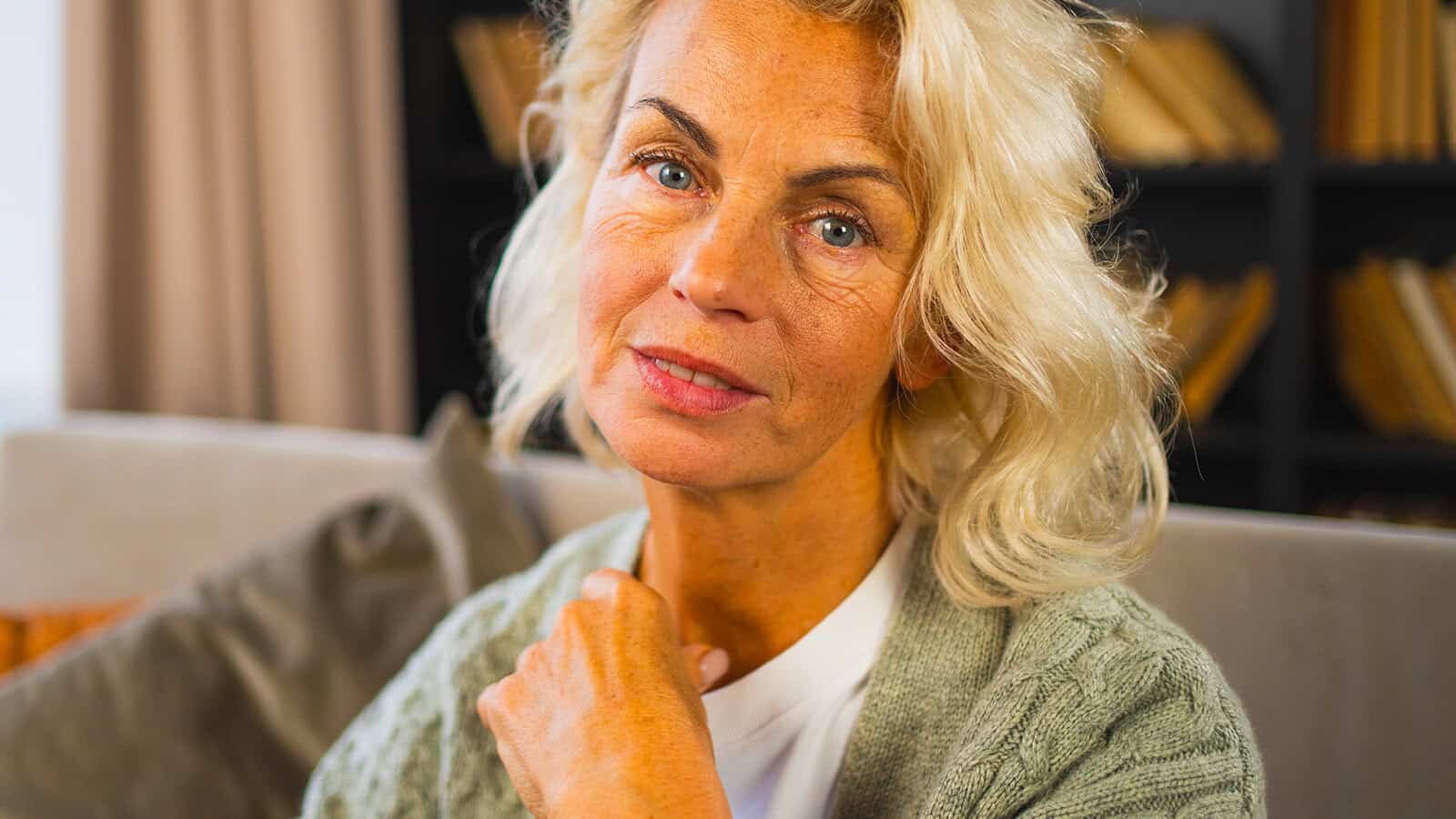 A woman with light blonde hair and blue eyes sits indoors, wearing a light gray cardigan and white shirt, looking thoughtfully at the camera. Bookshelves and a sofa are in the blurred background, suggesting a quiet moment of reflection—perhaps after a phone call that touched on themes of psychology or older adults’ loneliness.