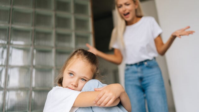 A young girl sits hugging her knees, looking sad, while a woman in the background appears to be yelling or arguing, arms raised in frustration.