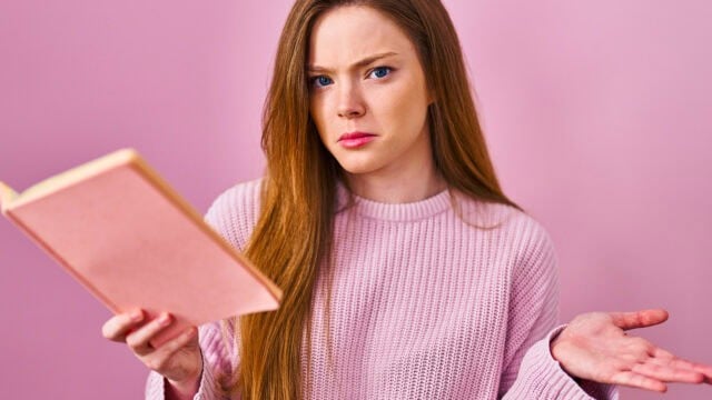 A woman with long red hair wearing a pink sweater holds an open book and looks confused, raising one hand in a questioning gesture, with a pink background.