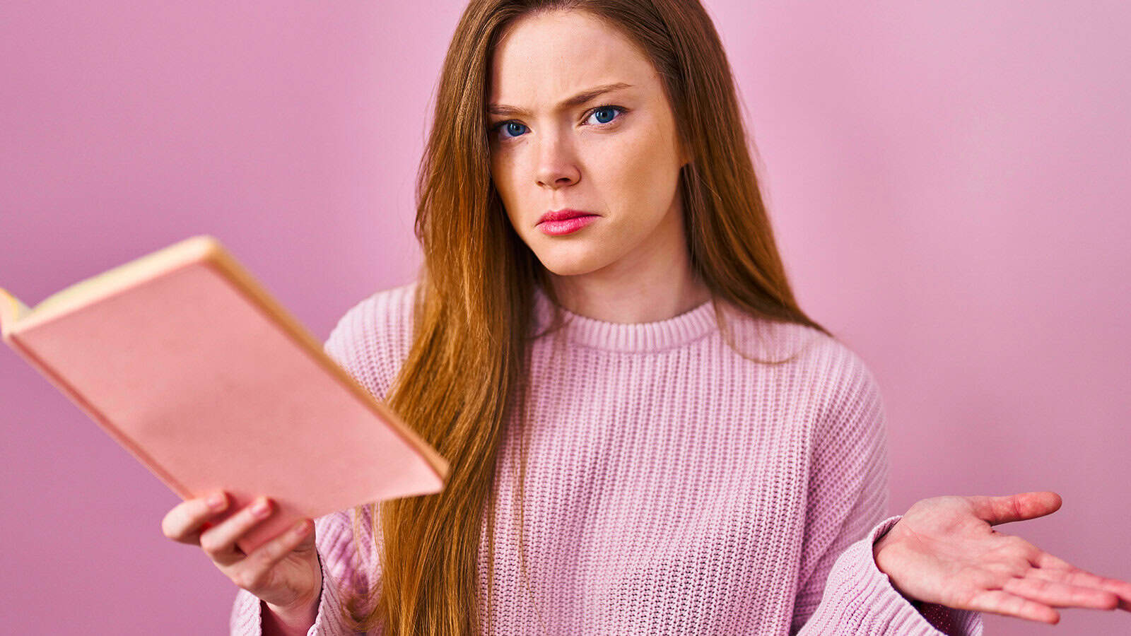 A woman with long red hair wearing a pink sweater holds an open book and looks confused, raising one hand in a questioning gesture, with a pink background.