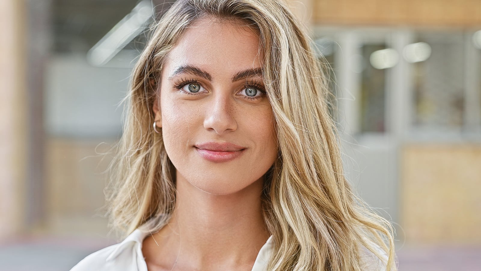 A young woman with long blonde hair and blue eyes smiles gently at the camera, standing outdoors with a blurred background of a building.