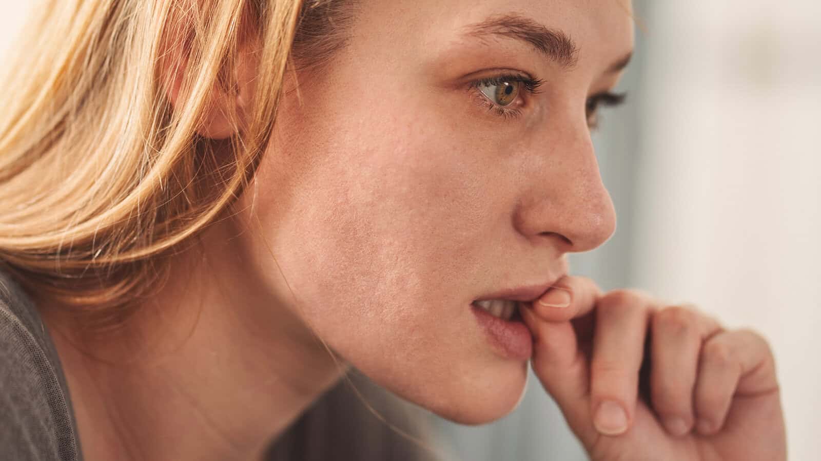 A person with light brown hair looks thoughtful and anxious, biting their fingernail while gazing into the distance.