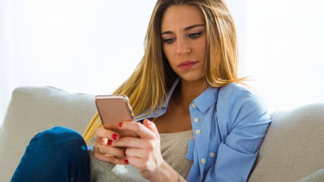 A woman with long blonde hair sits on a couch, looking thoughtfully at her smartphone. She wears a light blue shirt and casual jeans, with natural light coming from behind her.