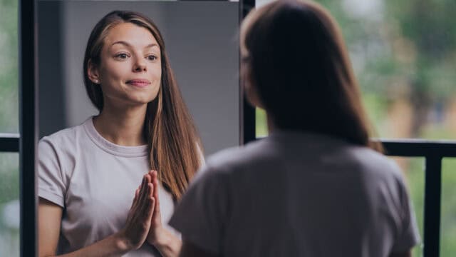 A woman with long brown hair stands in front of a mirror, hands pressed together in a thoughtful gesture, wearing a white t-shirt. Her reflection is visible, and she appears calm and reflective.