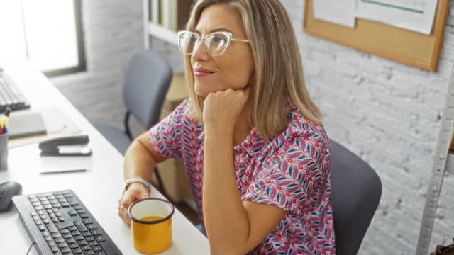 A woman with glasses sits at a desk, holding a yellow mug and looking thoughtfully to the side. She wears a colorful patterned shirt. A keyboard, computer, and notebook are on the desk.