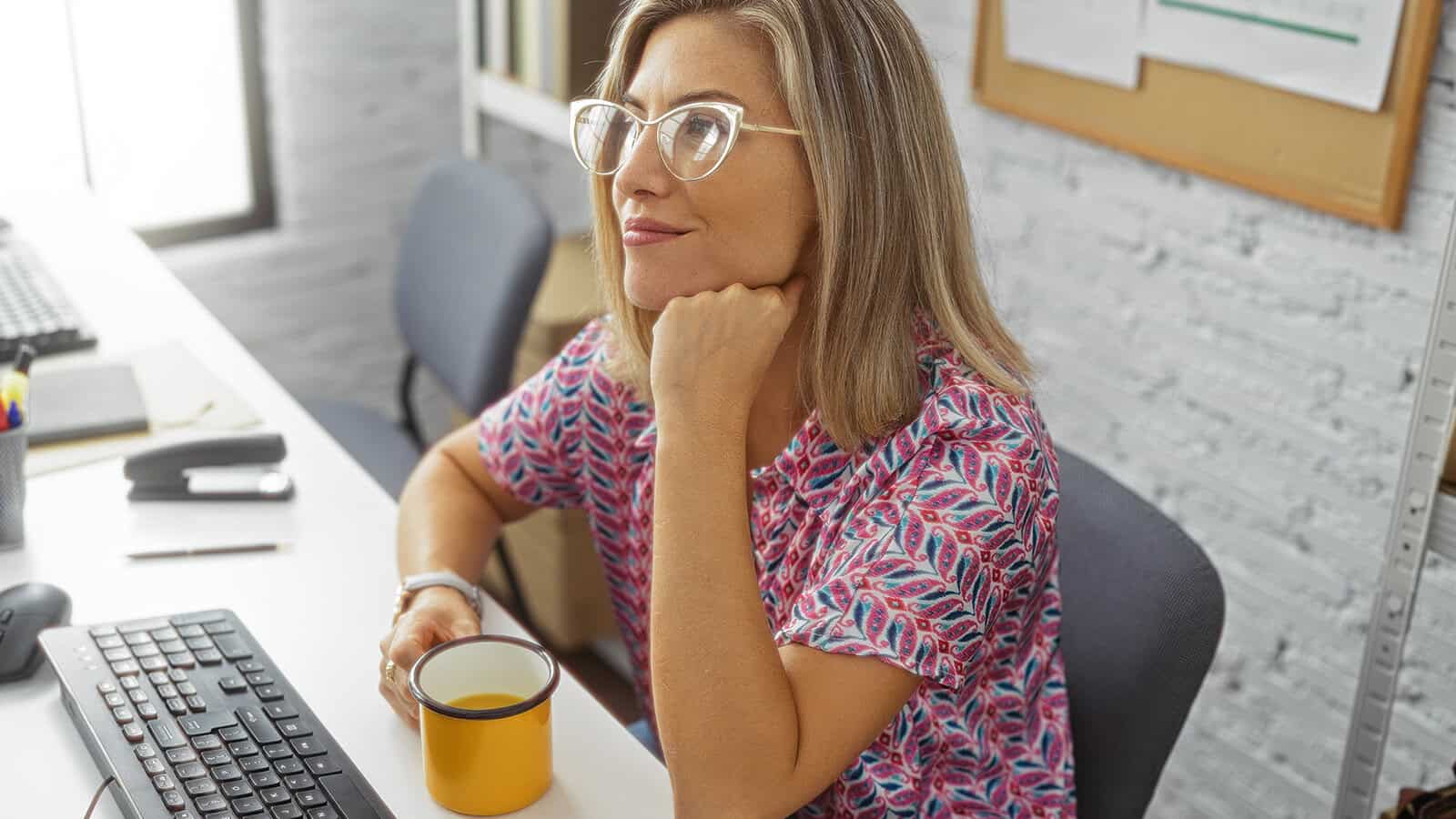 A woman with glasses sits at a desk, holding a yellow mug and looking thoughtfully to the side. She wears a colorful patterned shirt. A keyboard, computer, and notebook are on the desk.