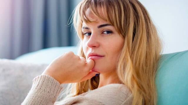 A woman with blonde hair and bangs sits indoors, resting her chin on her hand and looking thoughtfully at the camera. She wears a beige sweater and appears relaxed, with a blurred background behind her.