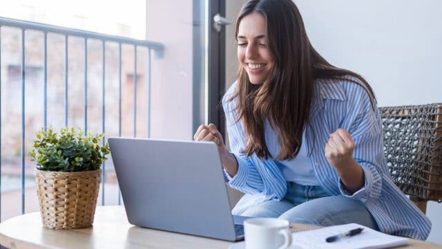 A woman sits at a table in front of a laptop, smiling and clenching her fists in excitement. A notebook, mug, and potted plant are on the table. She appears happy and engaged, with natural light coming through a window.