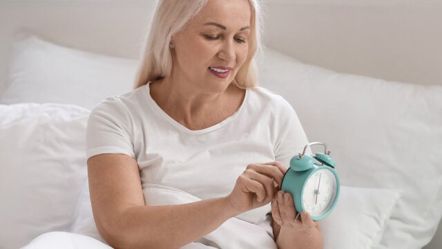 A woman with light hair sits on a bed, smiling as she adjusts a turquoise alarm clock. She wears a white shirt and is surrounded by white bedding.