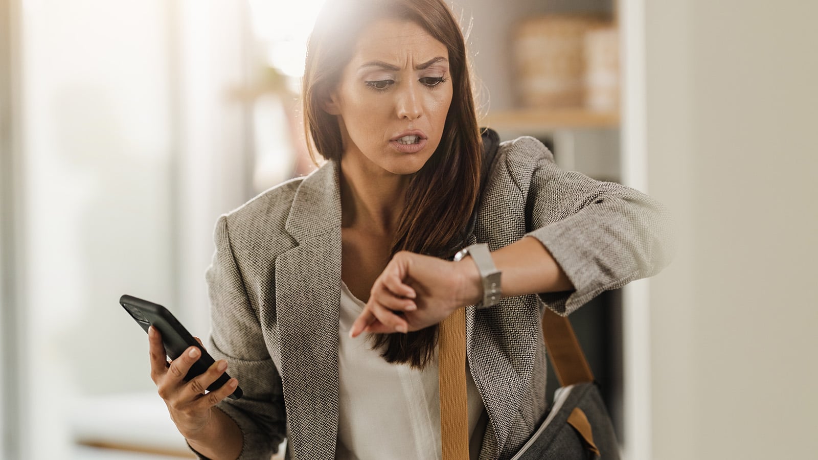 A woman holding a smartphone looks worriedly at her wristwatch, appearing constantly busy or late. Dressed in business attire with a bag over her shoulder, she stands indoors in a well-lit room, reflecting the psychology of self-avoidance.