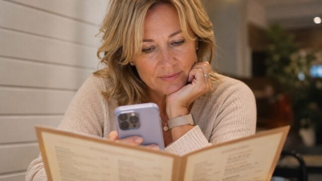 A woman sits at a table, looking at her smartphone in one hand while holding a menu in the other. She appears thoughtful, wearing a beige sweater, with short blond hair, in a warmly lit indoor setting.