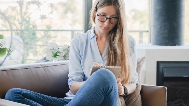 A woman with long hair and glasses sits on a couch, writing in a notebook. She is wearing a light blue shirt and jeans. The background shows large windows with natural light and some greenery outside.
