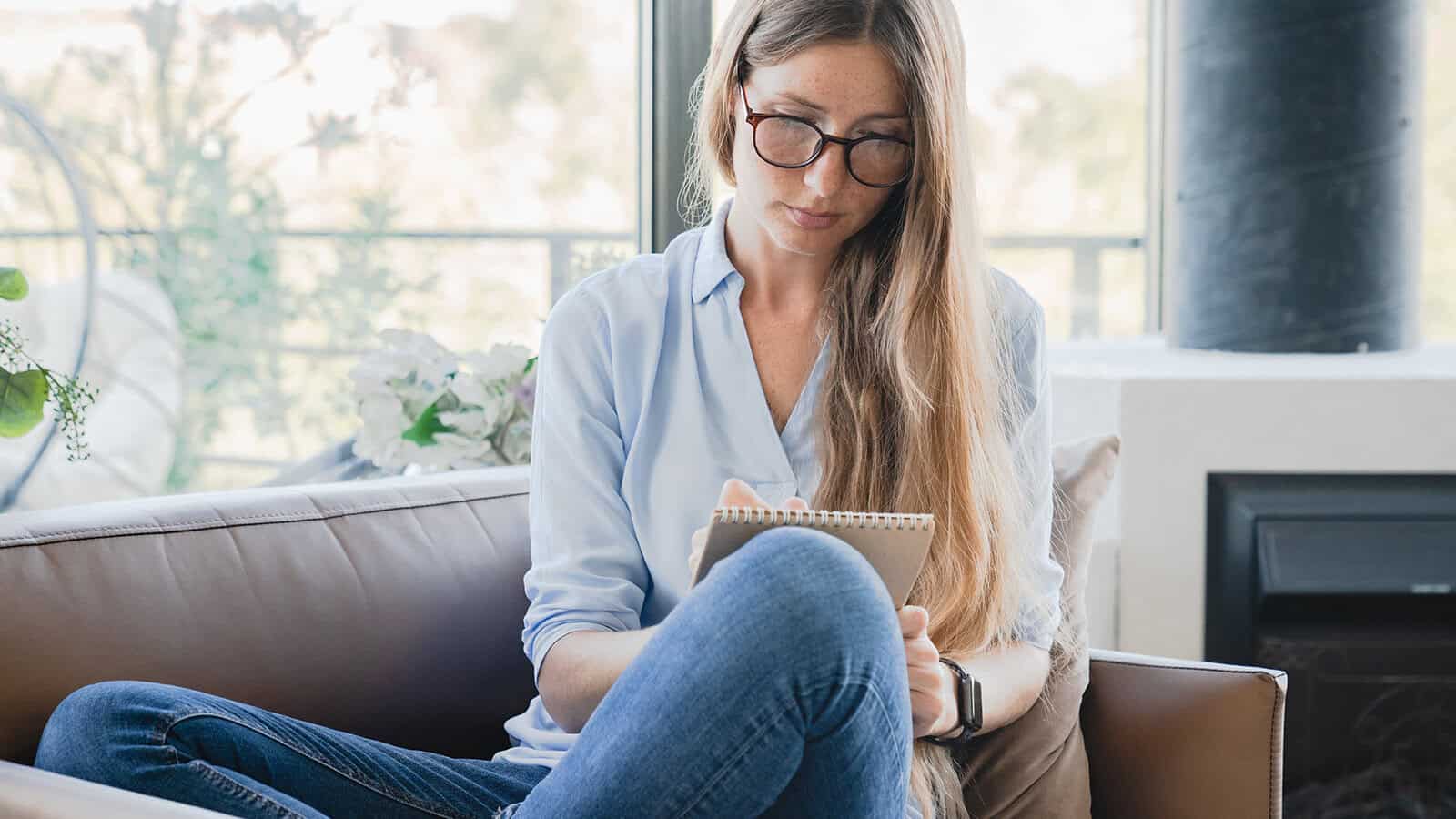 A woman with long hair and glasses sits on a couch, writing in a notebook. She is wearing a light blue shirt and jeans. The background shows large windows with natural light and some greenery outside.