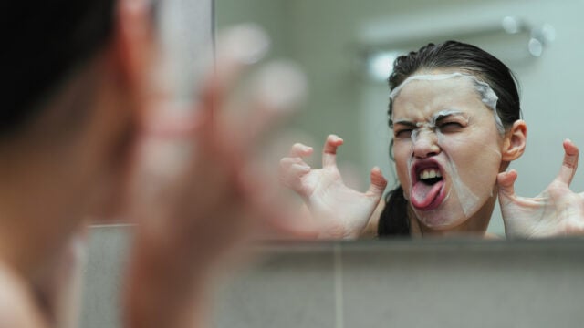 A person with a facial mask makes a funny face and claw gesture in front of a bathroom mirror, sticking out their tongue playfully.