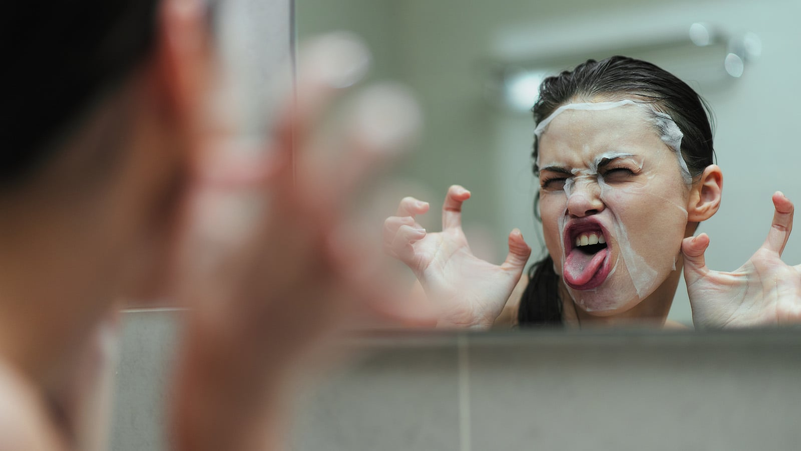 A person with a facial mask makes a funny face and claw gesture in front of a bathroom mirror, sticking out their tongue playfully.