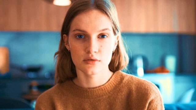 A young woman with light brown hair and a neutral expression sits indoors, wearing a tan sweater. The background is softly blurred, showing a kitchen setting with warm lighting.
