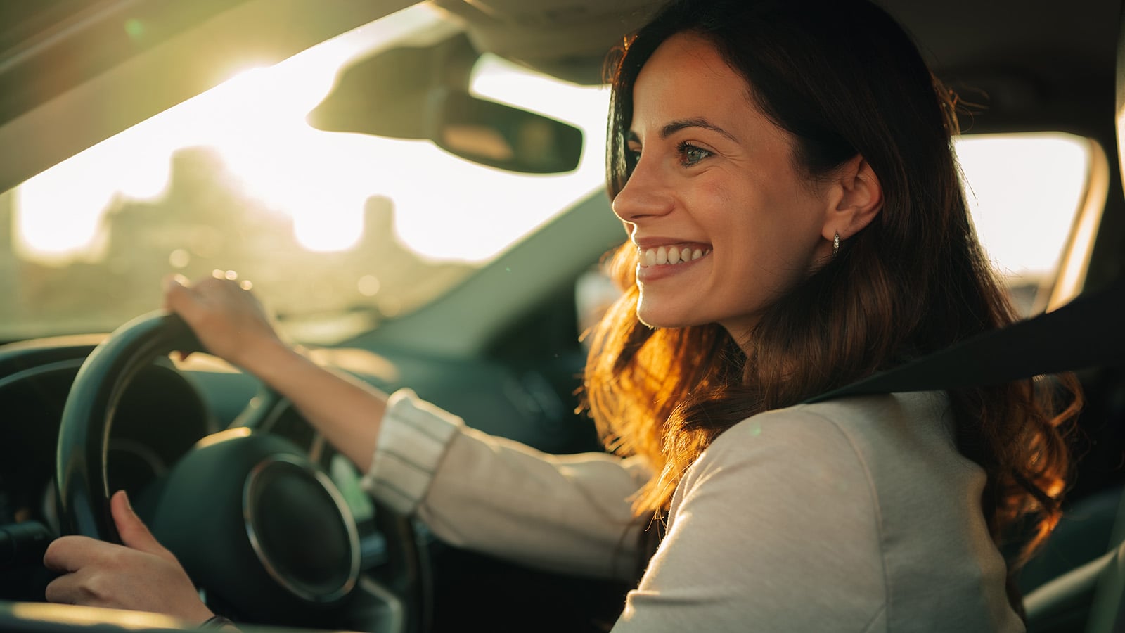 A woman with long brown hair smiles while driving a car during sunset, holding the steering wheel with one hand and looking forward.