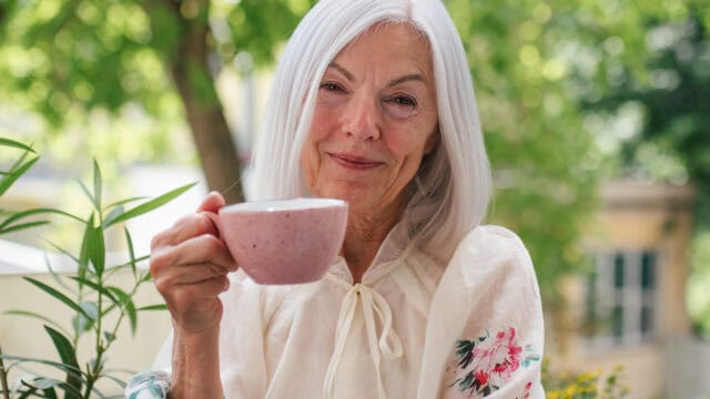 An older woman with gray hair and a floral blouse smiles while holding a pink mug outdoors, with green trees in the background.