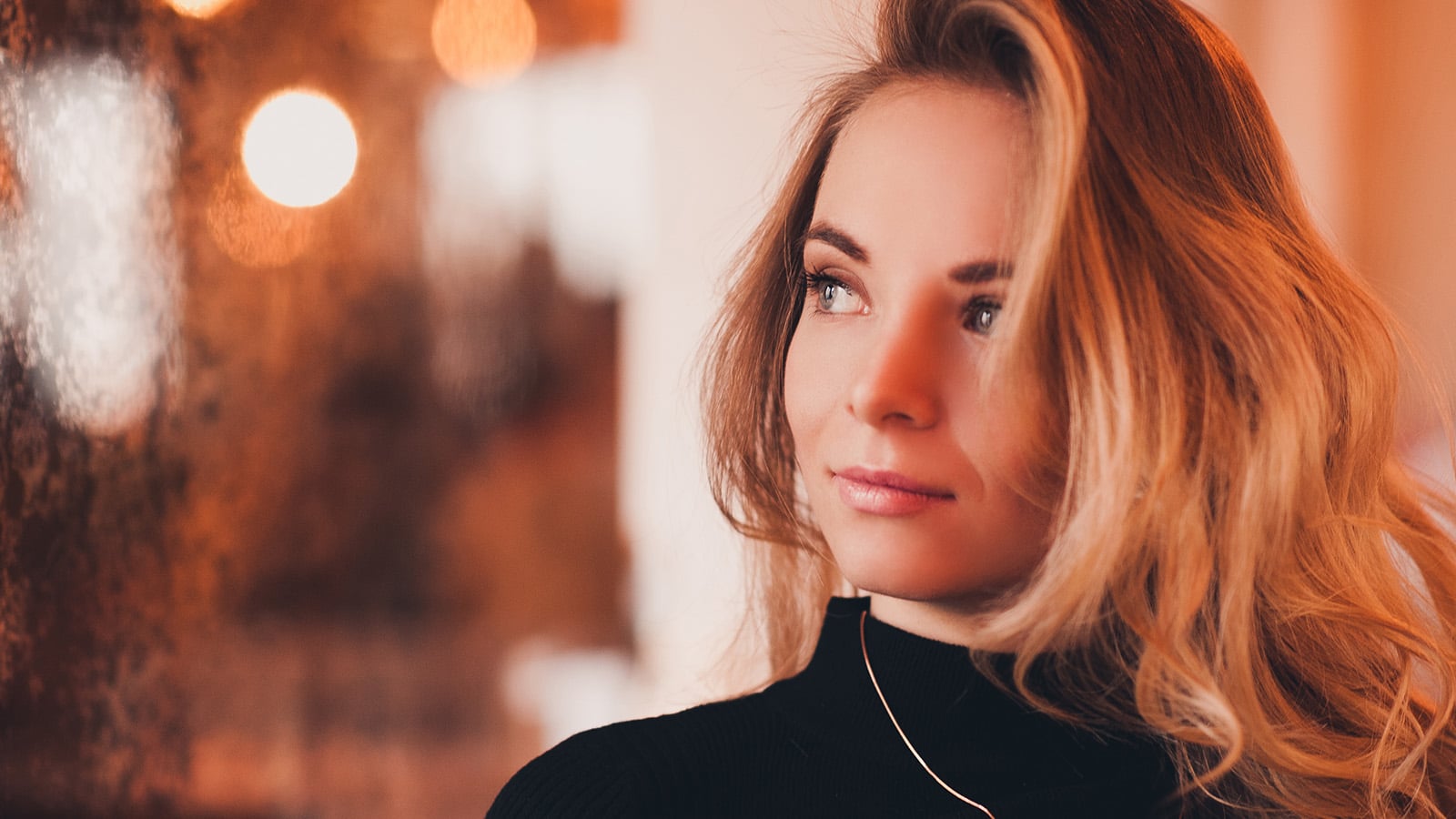 A woman with long blonde hair and a black turtleneck looks thoughtfully to the side. The background is softly lit with warm, blurry lights.