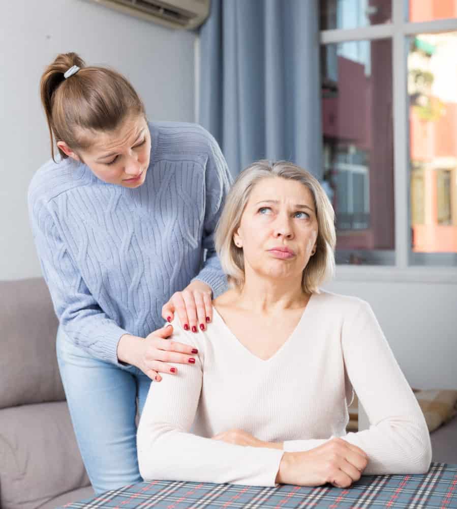A young woman stands behind an older woman, gently touching her shoulders. The older woman, experiencing the frustrations that can come with aging, looks away with a frustrated expression while the younger woman appears concerned—reflecting the challenges adult children often face in finding ways to show respect and support to their parents. They are indoors near a window.