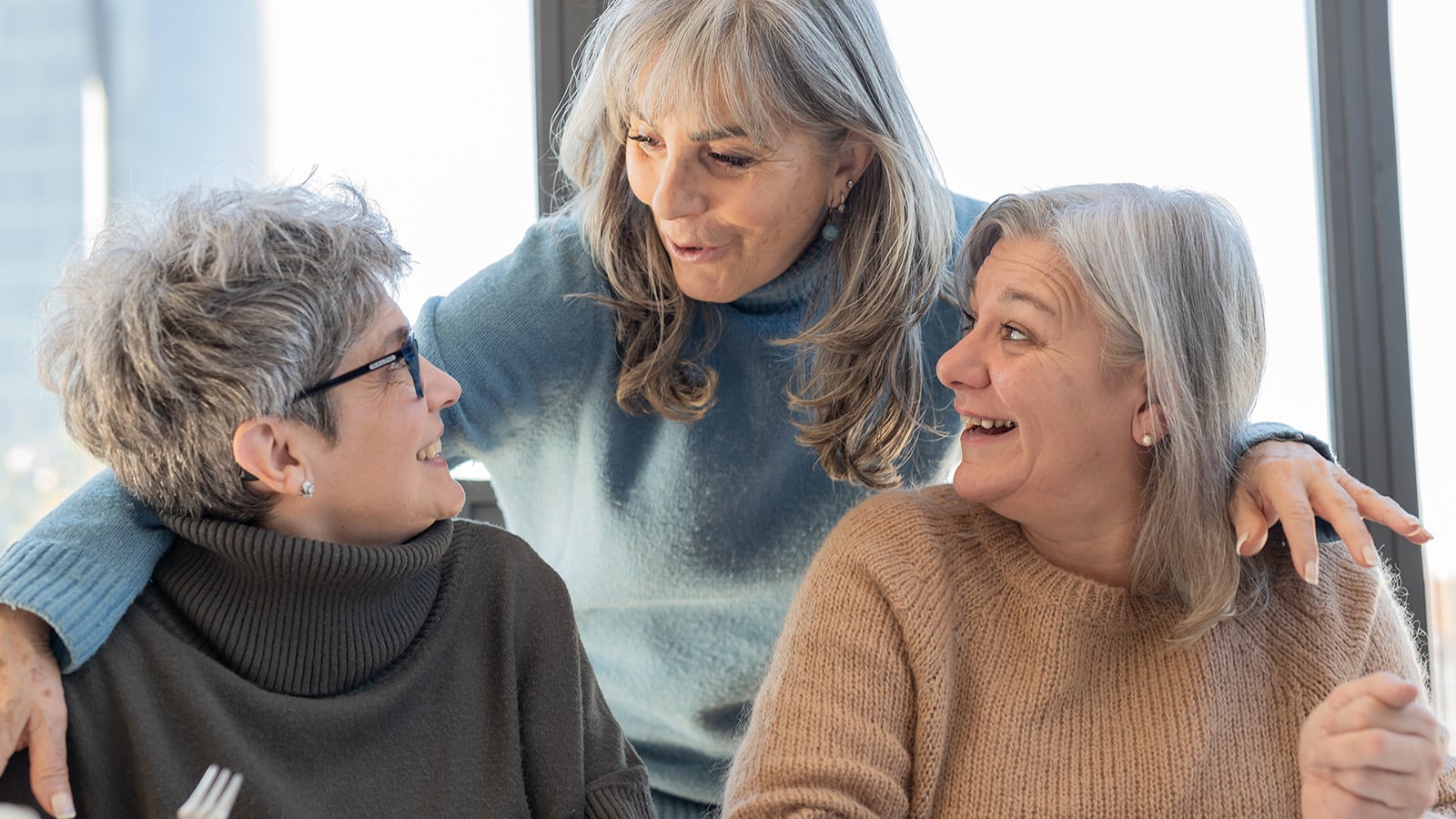 Three older women with gray hair are happily talking and smiling together indoors, with one standing and embracing the other two sitting at a table.