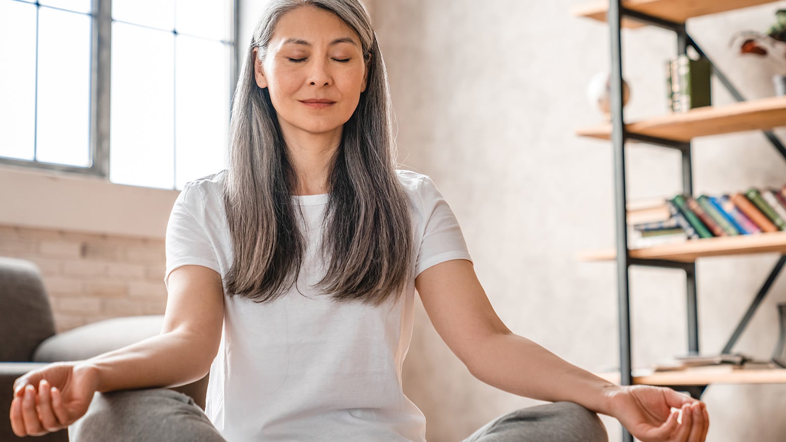 A woman with long gray hair sits cross-legged indoors, eyes closed, meditating in a peaceful room with sunlight, shelves, and books in the background.