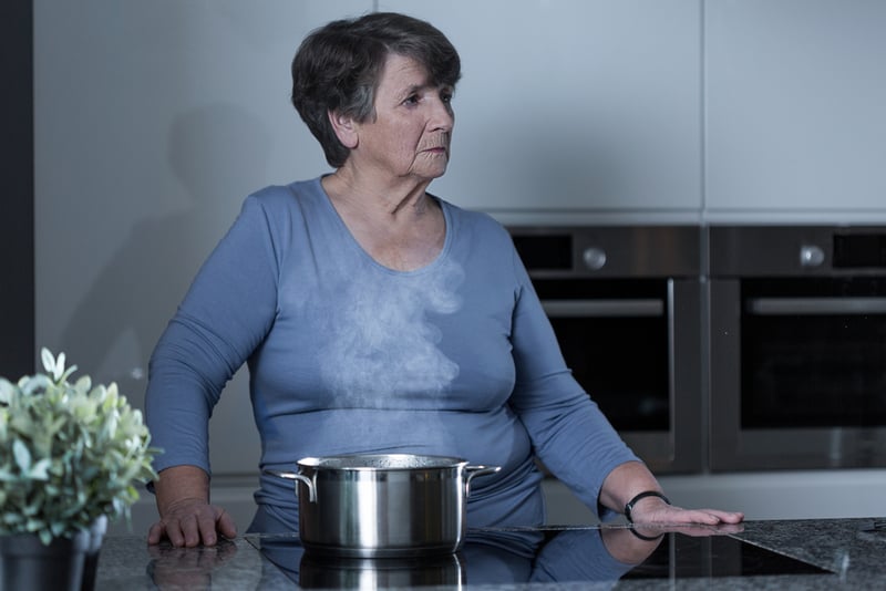 An older woman, a classic example of the boomer generation, sits in a blue shirt in her kitchen, looking thoughtfully to the side. A steaming pot rests on the glossy counter in front of her, and a green potted plant adds a touch of warmth nearby—perhaps suggesting that she’s deep in thought about communication and feelings shared around family meals.