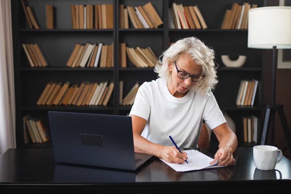 An older woman with gray hair and glasses writes on paper at a desk with a laptop and coffee mug, in front of a bookshelf filled with books—a quiet scene that reflects the thoughtful habits of people raised in the 60s and 70s, and hints at some of the things people are proud of across generational differences.