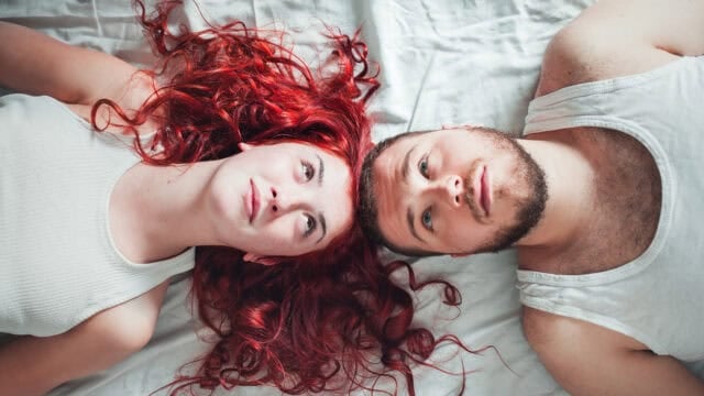 A woman with long, curly red hair and a man with a beard lie side by side on white sheets, both wearing white tank tops, looking upward with relaxed expressions.