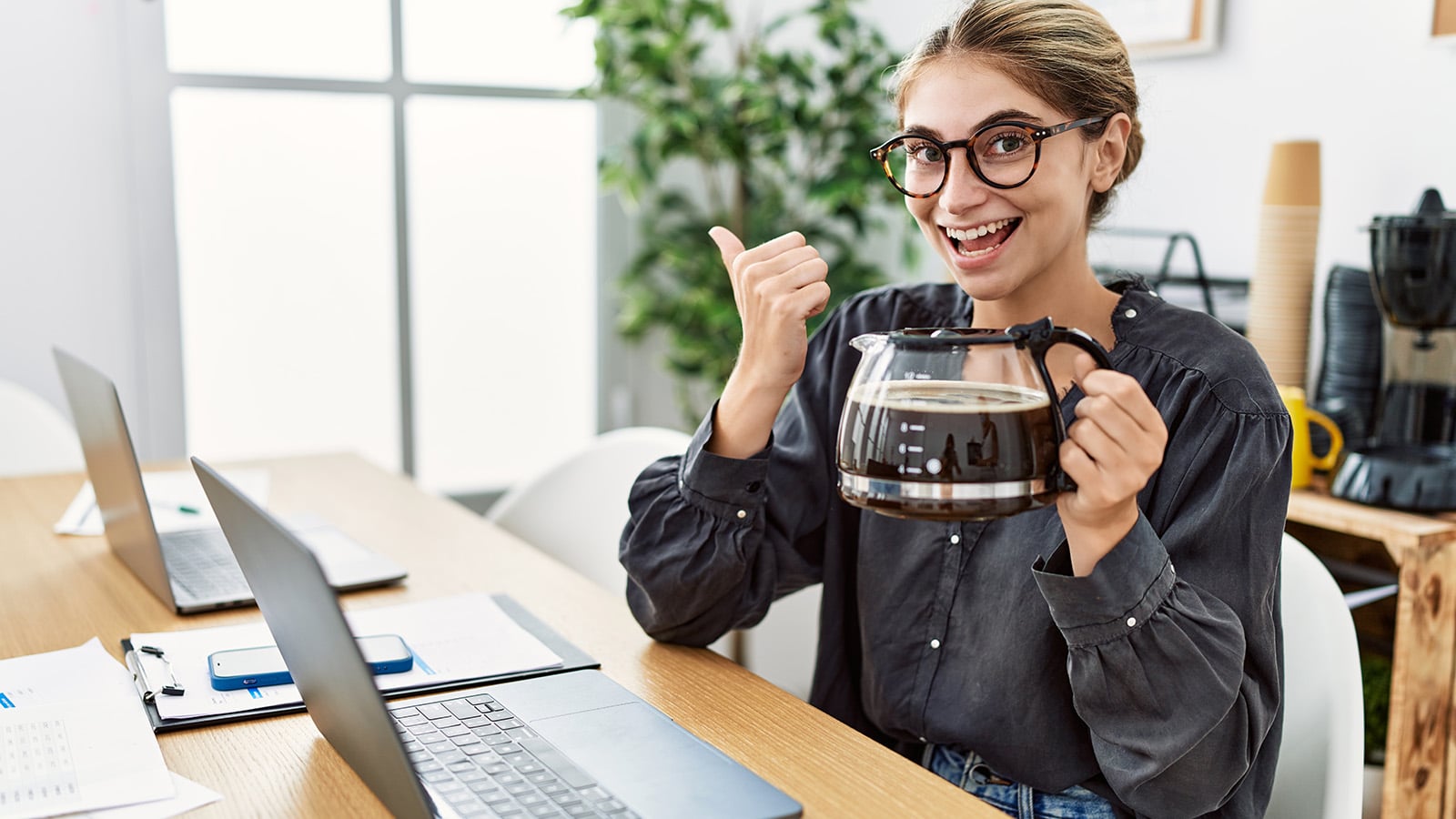 A smiling person wearing glasses sits at a desk with laptops, holding a large coffee pot in one hand and giving a thumbs up with the other. The workspace has plants and coffee supplies in the background.