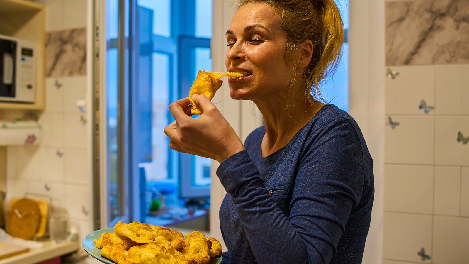 A woman in a blue shirt enjoys eating fried food from a plate while standing in a kitchen with white tiles and an open window in the background.
