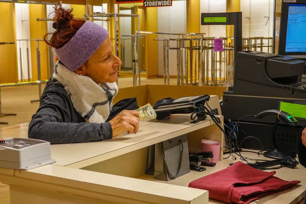 A woman with a purple headband, reminiscent of styles popular in the 60s and 70s, holds cash while speaking to a cashier at a retail counter. Behind her, empty clothing racks and a STOREWIDE! sale sign are visible.