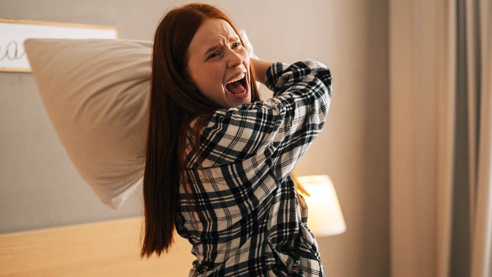 A person with long red hair in plaid pajamas smiles widely and playfully swings a pillow in a cozy, softly lit bedroom.