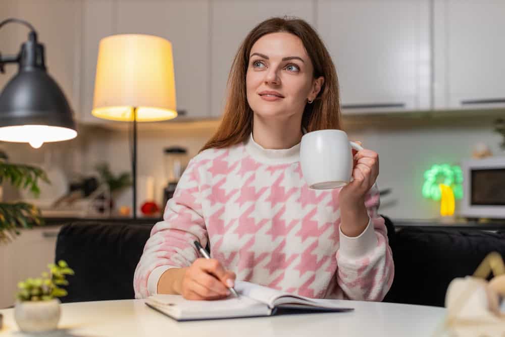 A woman in a pink and white sweater sits at a table, holding a white mug and writing in her notebook, engaging in daily journaling. She looks off to the side, appearing thoughtful—as if immersed in the process of life processing. The background shows a cozy, well-lit kitchen.