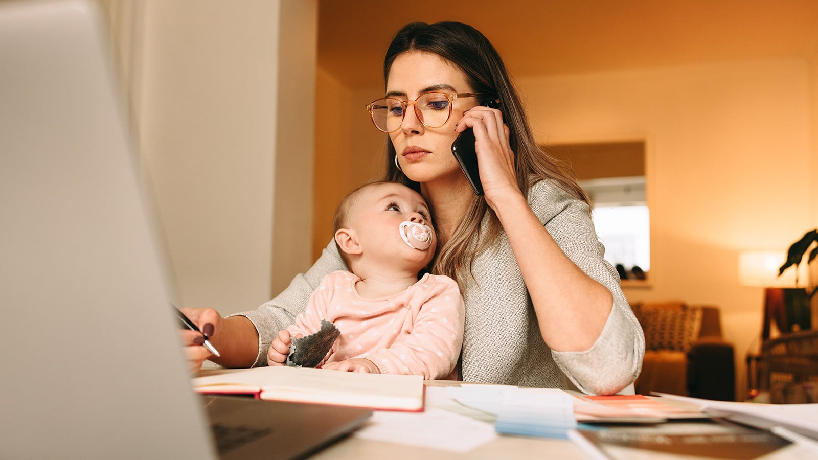 A woman working at a desk with a laptop, holding a baby in one arm and talking on the phone, surrounded by papers and notebooks.