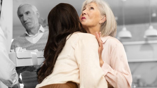 Two women hug; one looks unenthusiastic while a man in the background watches and holds a gift box. The background is grayscale, while the people are in color.
