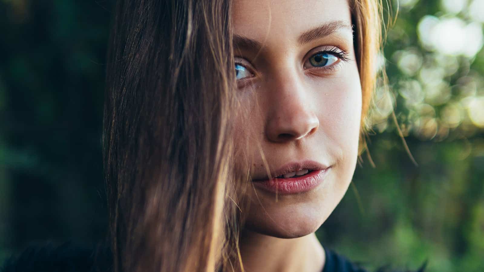 A close-up of a woman with long brown hair partly covering her face, blue eyes, and a neutral expression, standing outdoors with a blurred green background.