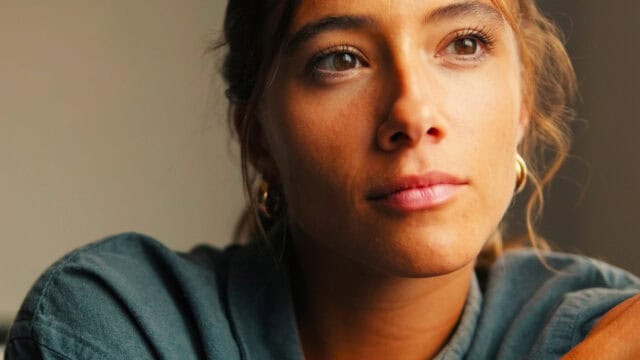 A person with light brown hair and gold hoop earrings gazes thoughtfully into the distance, softly lit by natural light, wearing a blue-green collared shirt.