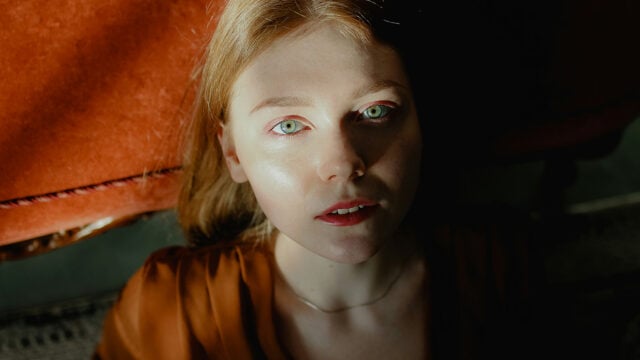 A person with fair skin and light hair looks up toward soft light, casting shadows on their face. They wear a brown top and are seated near an orange surface, creating a dramatic, contemplative mood.