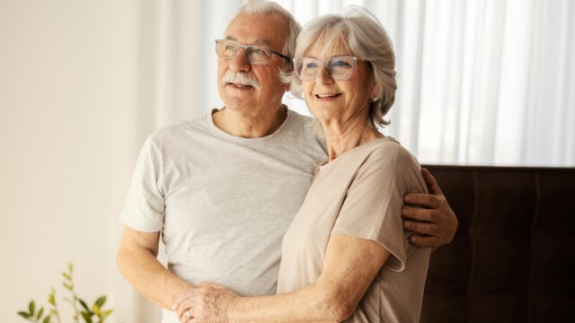 An elderly couple with gray hair and glasses stand close together, smiling and looking off into the distance, capturing the warmth of boomer parents enjoying family relationships in a bright, softly lit room with white curtains in the background.