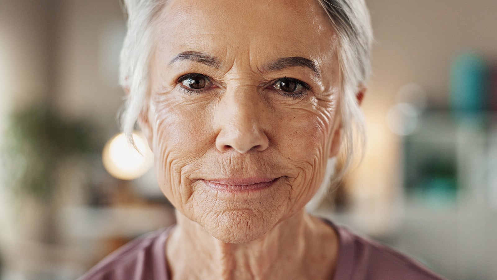 Close-up of an older woman with gray hair and wrinkled skin, wearing a light purple top, looking directly at the camera with a subtle, gentle smile—a graceful presence that reflects the wisdom and resilience often admired among boomers. The background is softly blurred.